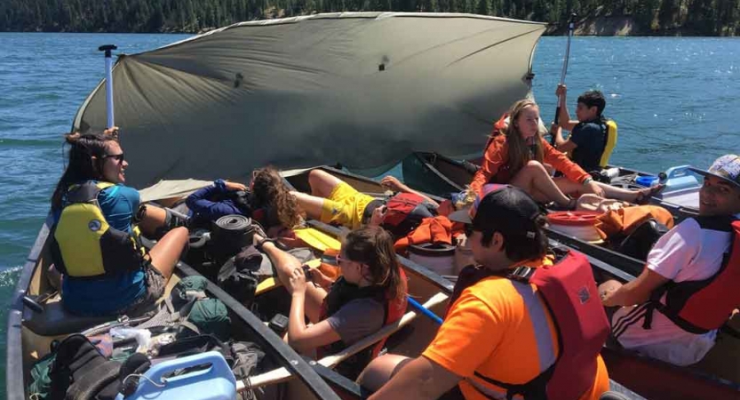 a group of canoes tethered together hold high school students as they use their paddles to prop up a tarp for shade